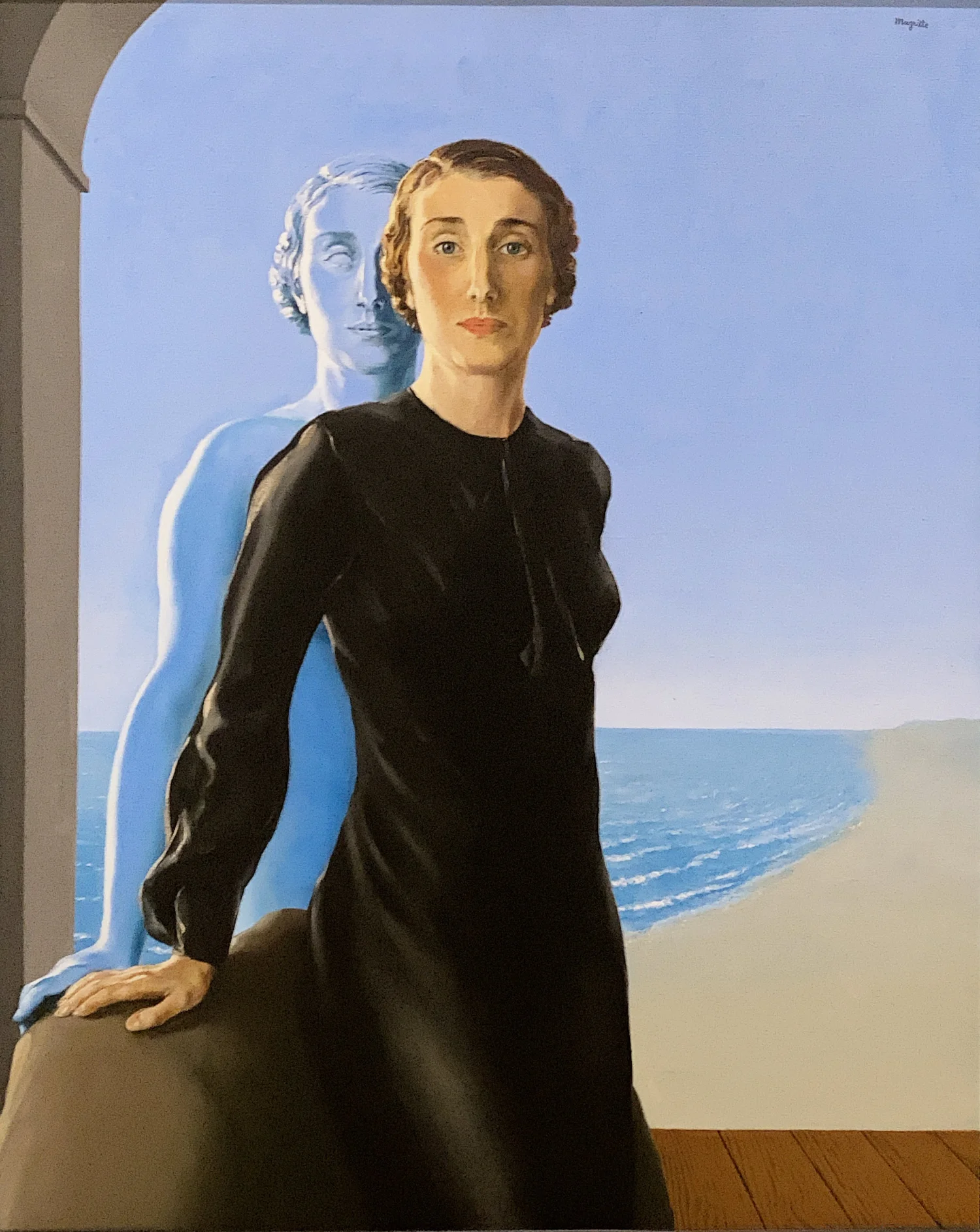 A woman in a black dress standing in front of a mural of a blue-toned bust of a woman, with a beach scene in the background.