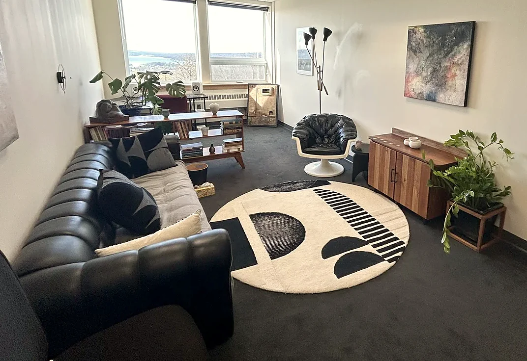 The consulting room at 950 Danby Road in Ithaca: a leather couch, bookshelf, lounge chair and round rug, with windows looking out over the lake.